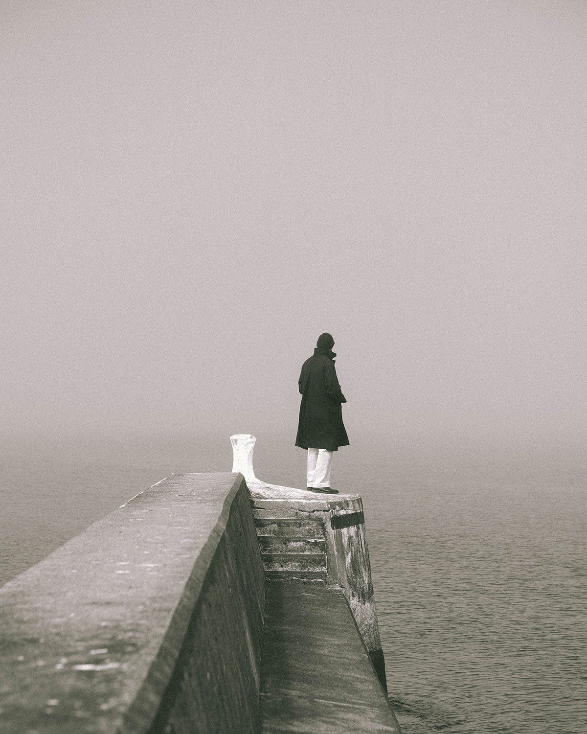A man in a dark coat and white trousers at the end of a stone pier, looking out over the water in fog