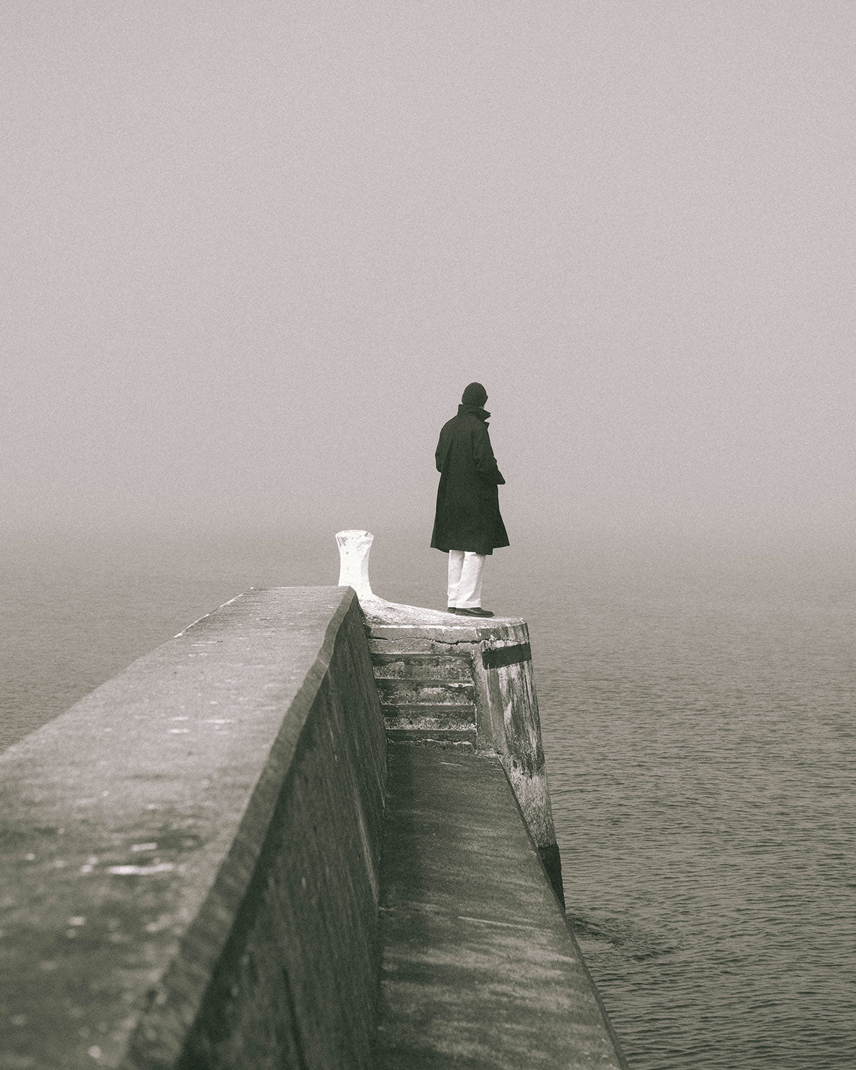 A man in a dark coat standing at the end of a stone pier by a white bollard, the sea lost in fog