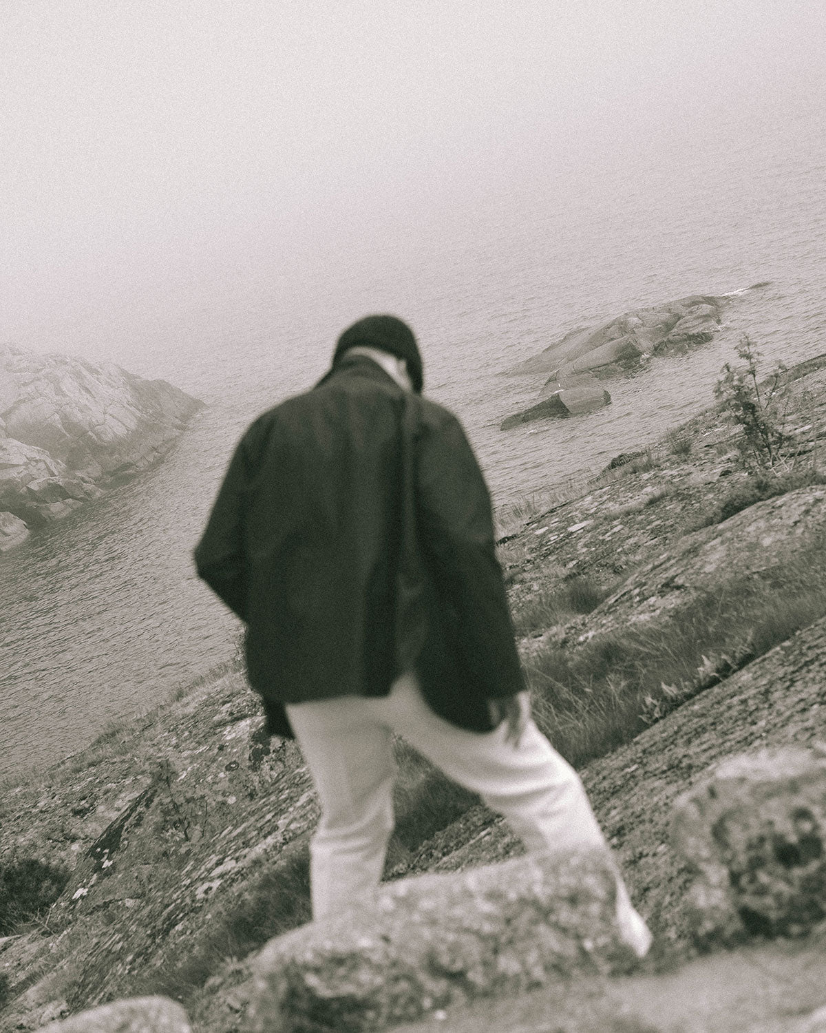 A man in a dark jacket and knitted hat climbing over coastal rocks, the sea behind in fog