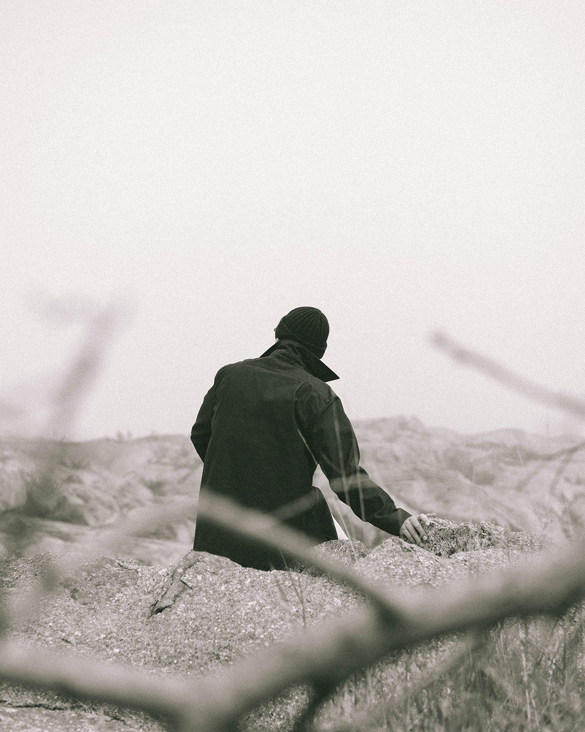 A man in a dark jacket and knitted hat seen from behind, a hand on the rock, through blurred grass
