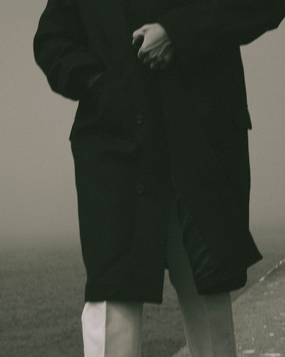 Detail of a dark coat and white trousers, a hand at the pocket, water behind