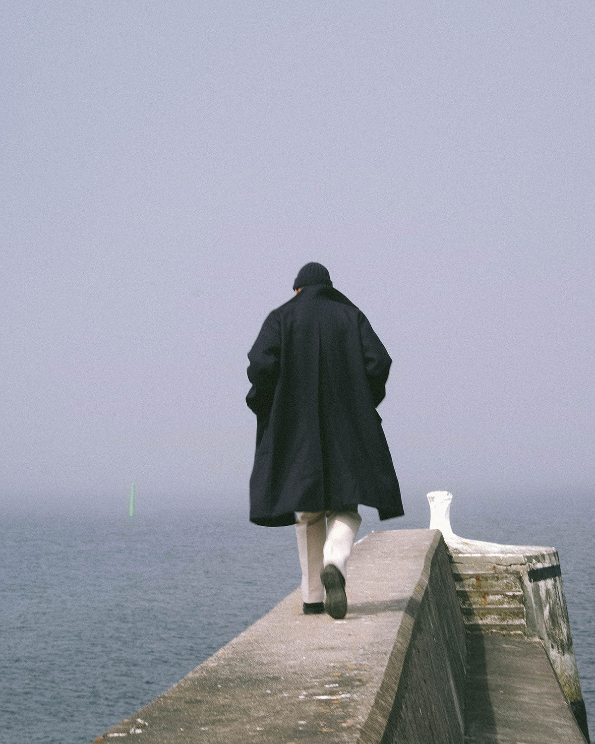 A man in a navy coat and knitted hat walking out along a stone pier towards a green marker in the fog
