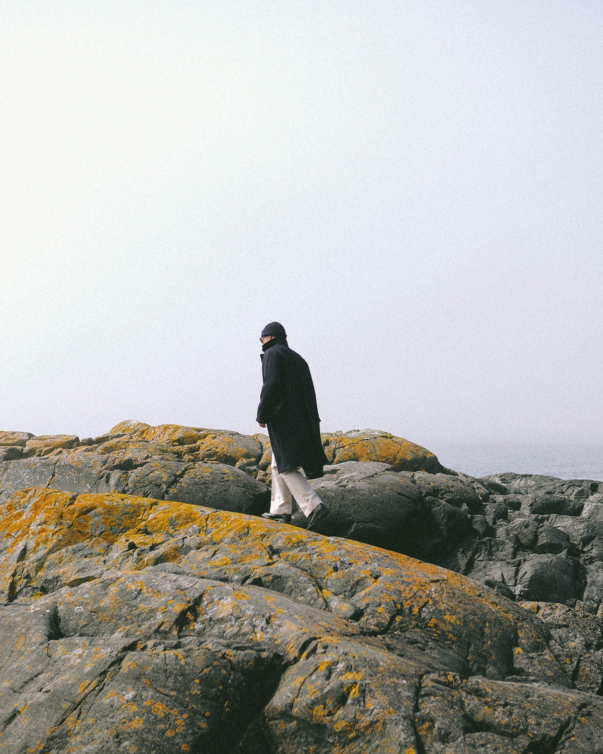 A man in a navy coat walking across rocks covered in orange lichen under a white sky