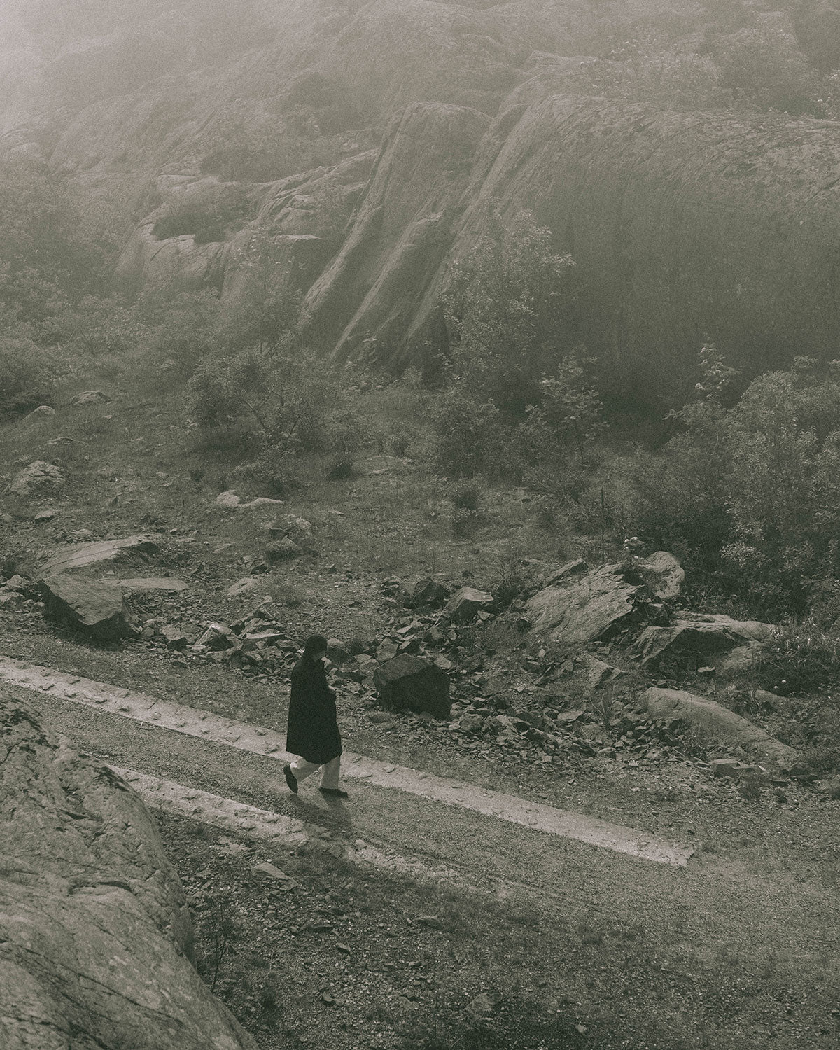 A man in a dark coat walking a path below rock faces in fog, seen from above